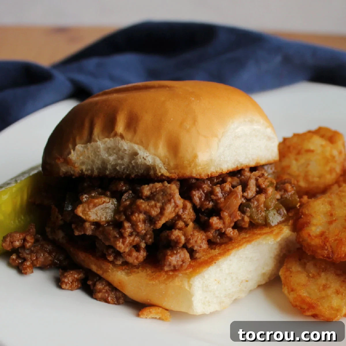 Cola Sloppy Joes Dinner Plate Toasted bun generously filled with crumbly cola sloppy joe mixture, served alongside crispy tater tots and a crunchy pickle spear.