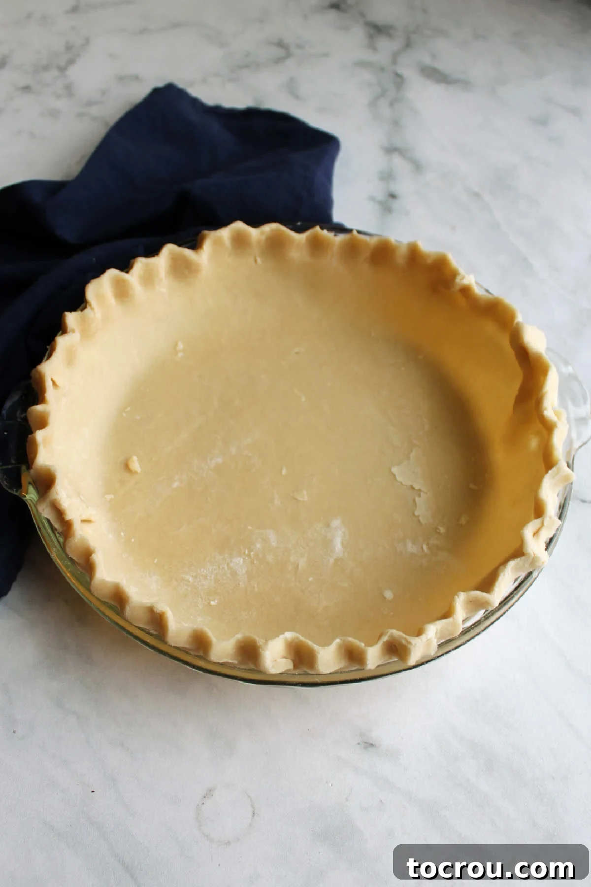 A pristine lard pie crust, perfectly shaped and with beautifully crimped edges, awaits its filling in a pie pan.