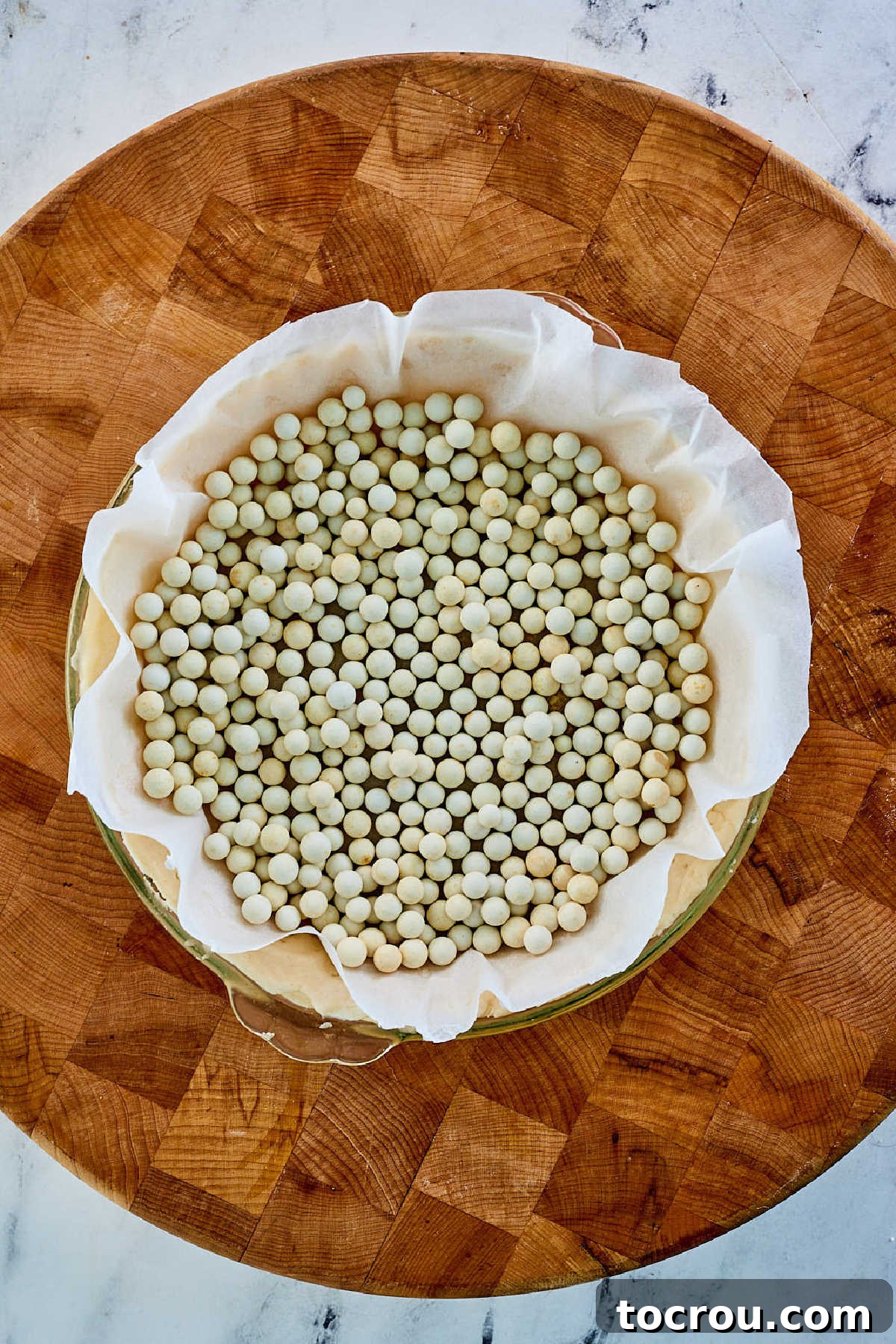 A glass pie plate containing a pre-baked pie crust, lined with parchment paper and filled with ceramic pie weights, ready for blind baking in the oven.