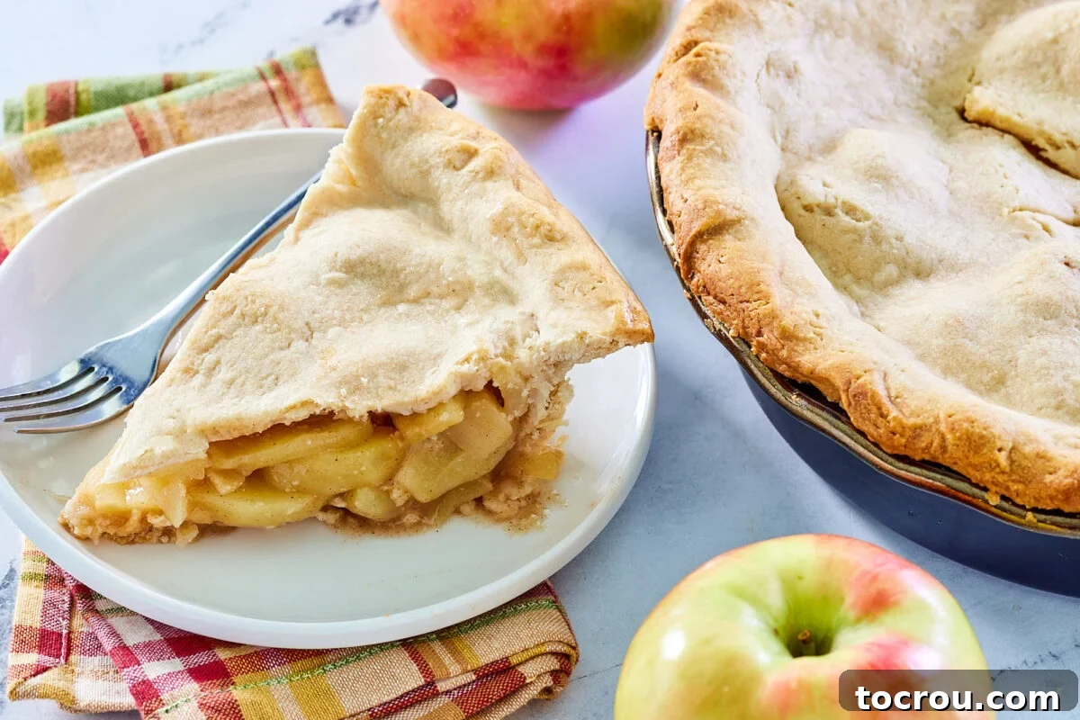Piece of grandma's deep dish apple pie next to a couple of fresh apples and the rest of the pie, showcasing its generous filling and golden crust.