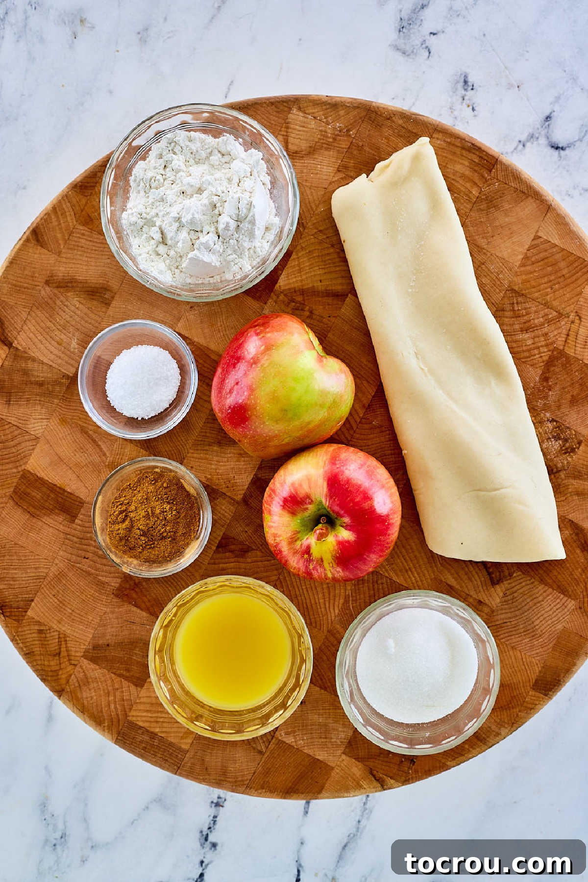 Ingredients for a deep dish apple pie laid out, including vibrant apples, a bowl of orange juice, sugar, cinnamon, flour, and a prepared pie pastry.