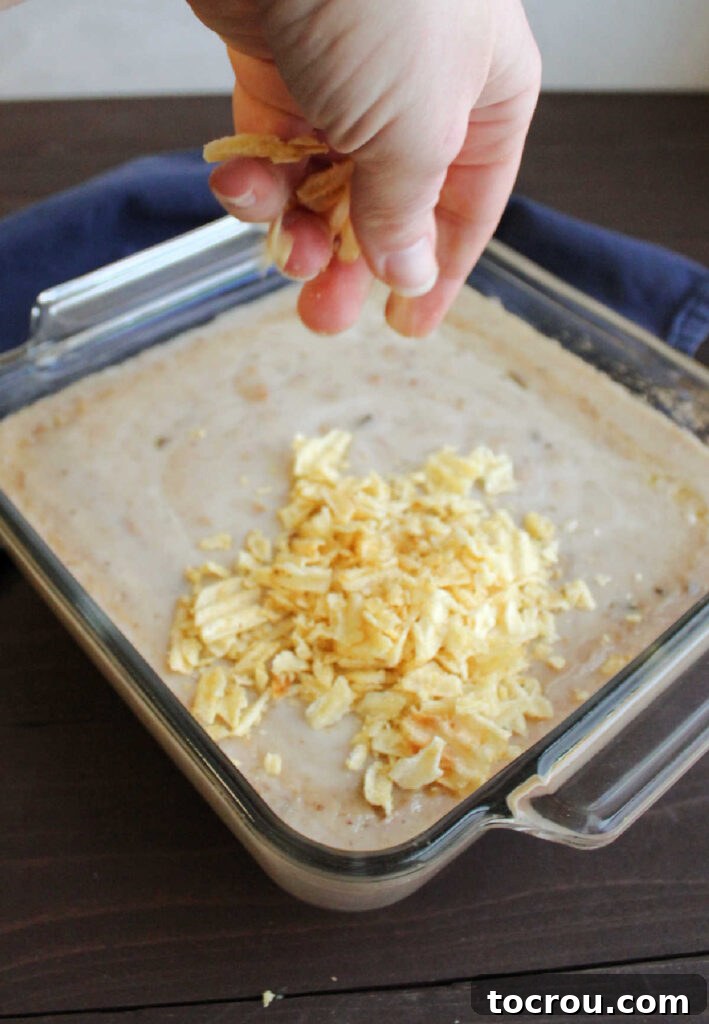 Mimi's Signature Tuna Bake 4 A hand gently sprinkling crushed potato chips over the bubbling, creamy tuna mixture in a baking dish, preparing for the final crispy topping.