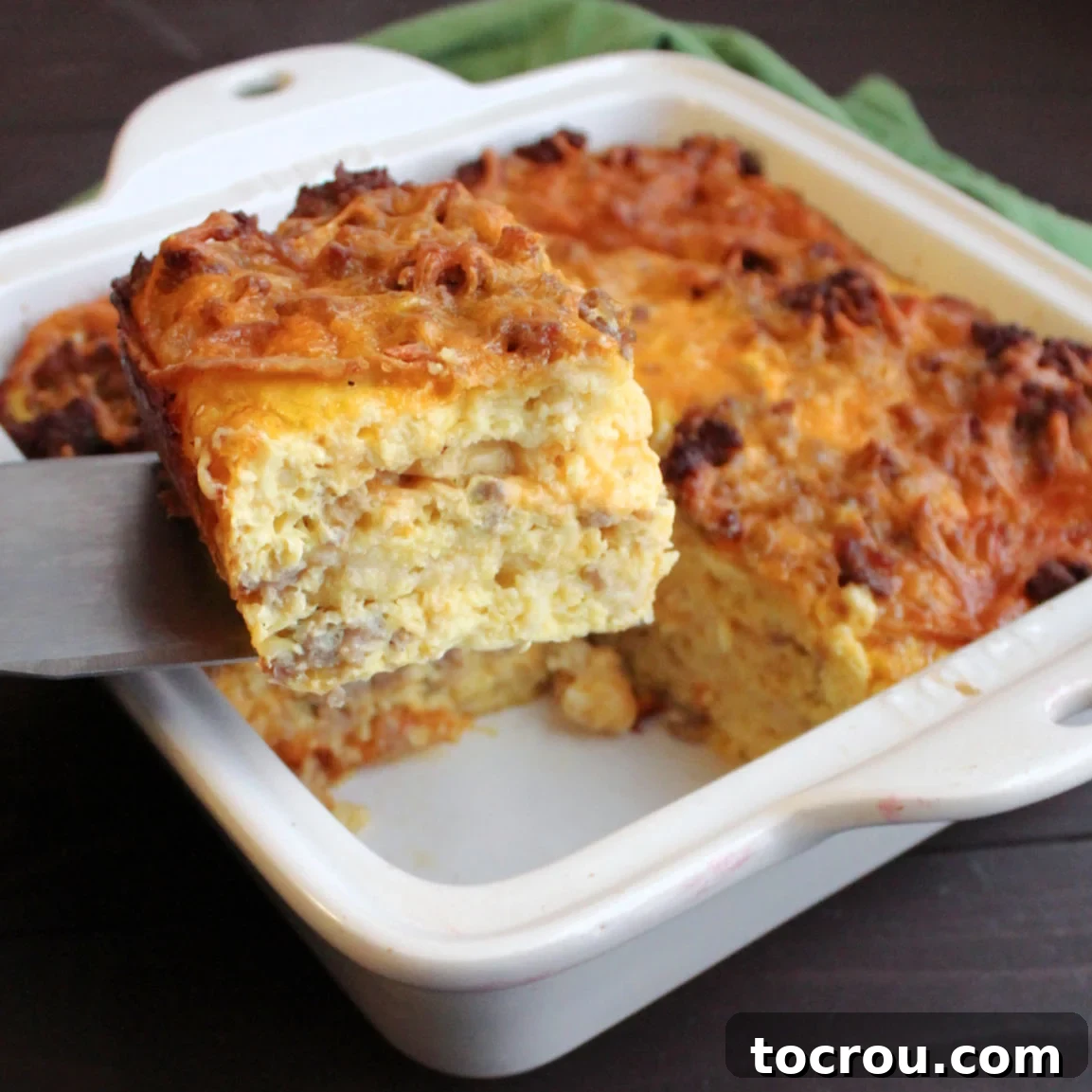 Sausage and Waffle Breakfast Casserole A square piece of golden-brown sausage and waffle breakfast casserole being lifted out of a baking dish, showing layers of waffle, egg, sausage, and cheese.