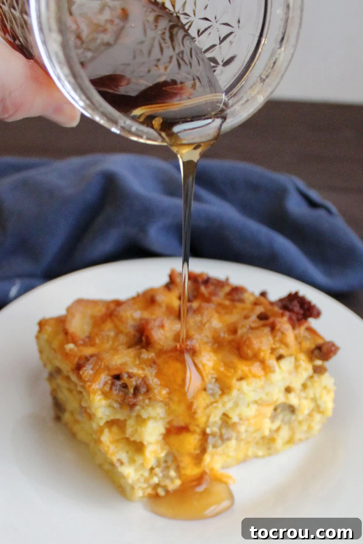 Serving Waffle Casserole with Syrup Maple syrup being poured generously over a golden-brown piece of waffle, egg, and sausage breakfast casserole on a white plate.