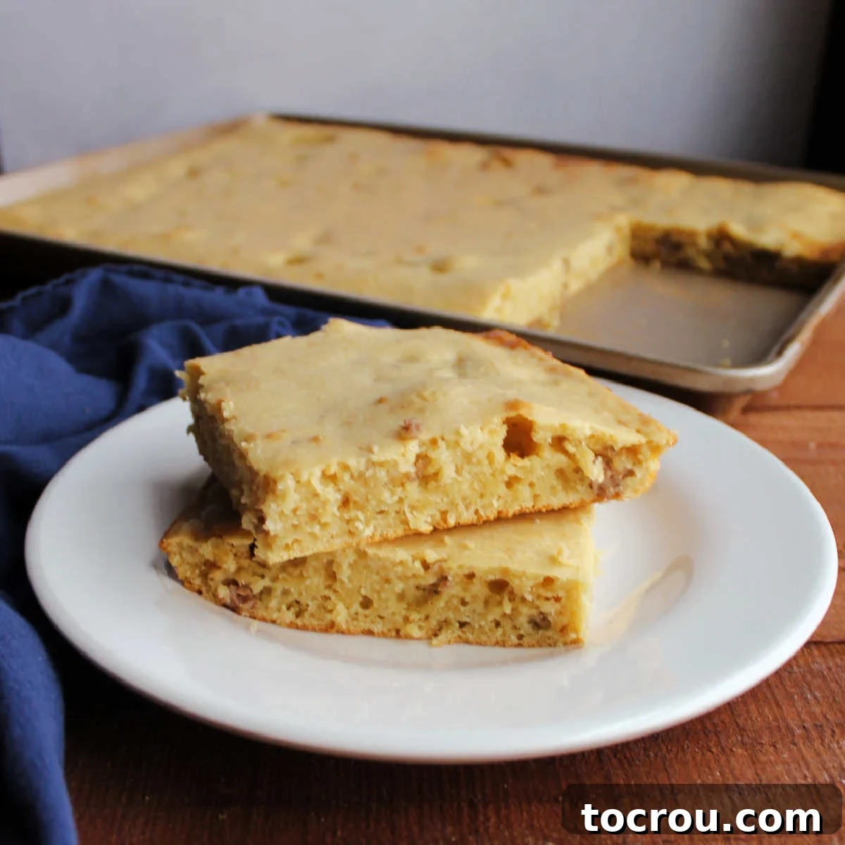 stack of two square slices of sheet pan pancake with chunks of breakfast sausage baked inside, drizzled with maple syrup.