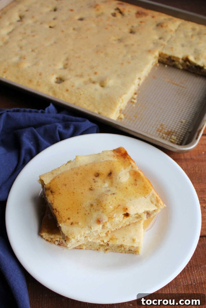 stack of pancake slices on plate with maple syrup, with the remaining sheet pan filled with pancake in the background.