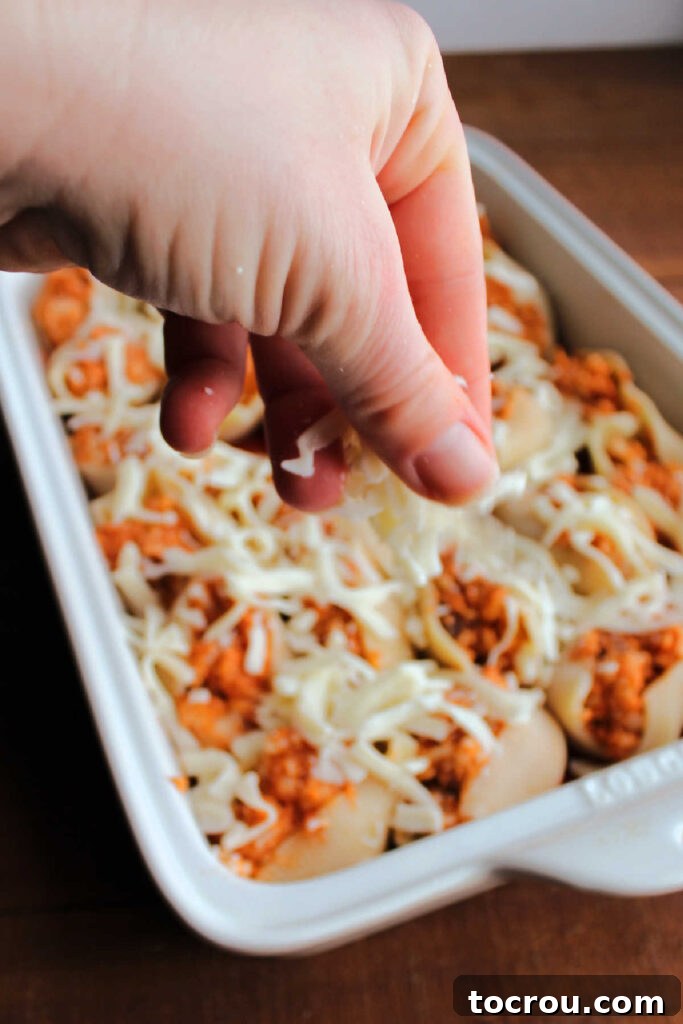 Hand sprinkling cheese over a pan of chicken Parmesan stuffed shells, almost ready to bake.