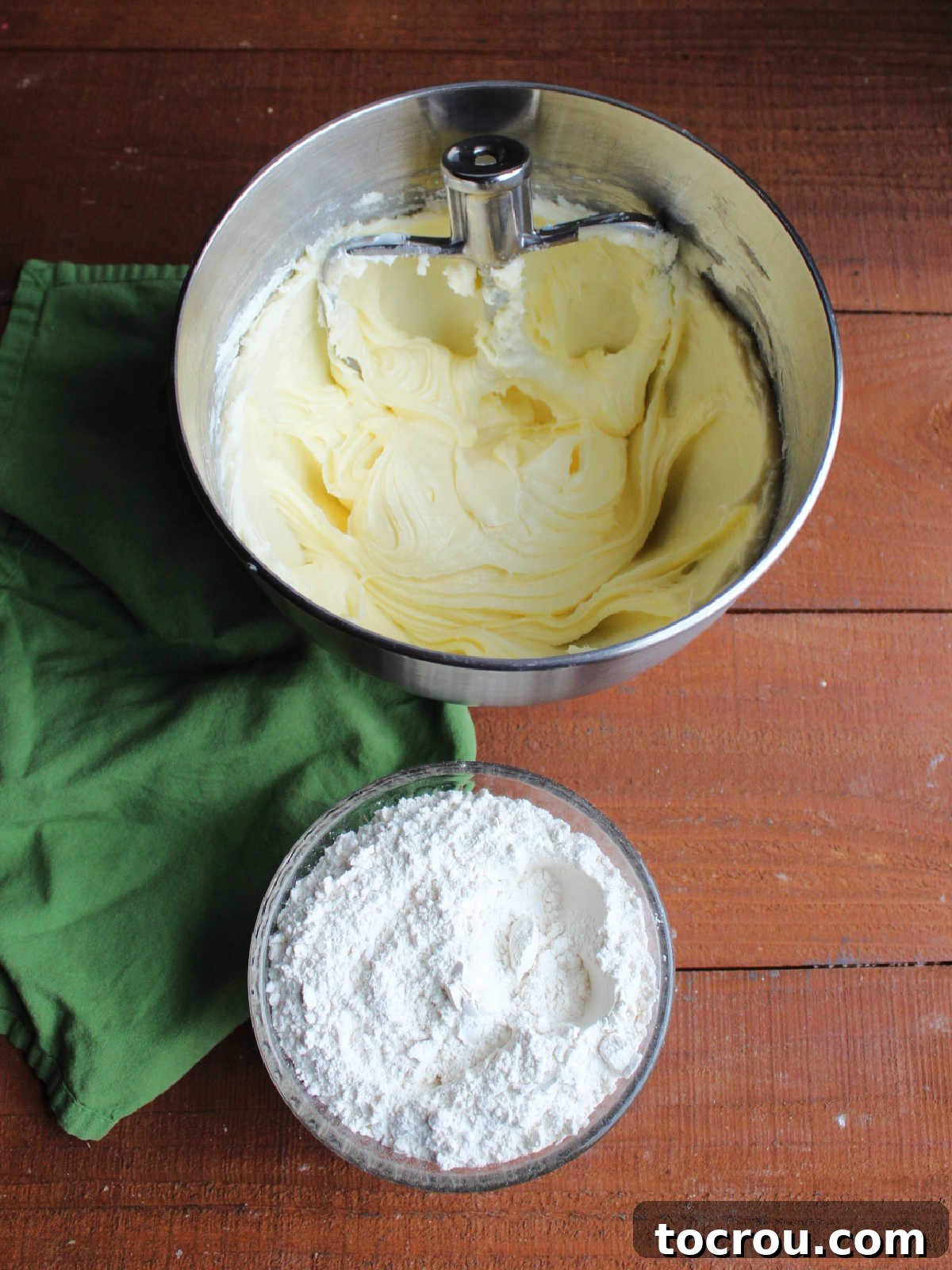 Mixer bowl with creamed butter, cream cheese, and sugar next to a bowl of dry ingredients, ready for combination to form sugar cookie dough.