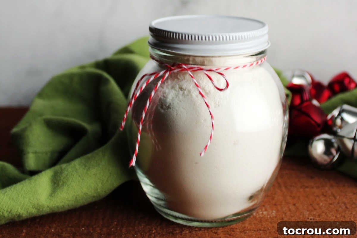 Small glass jar filled with homemade white hot chocolate mix, adorned with red and white twine, placed next to festive bells, evoking a sense of cozy winter comfort.