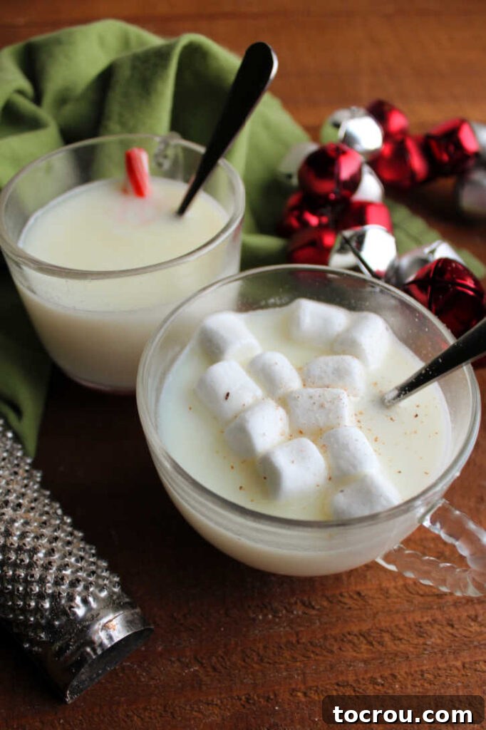 Two clear glass mugs of white hot chocolate: one garnished with a candy cane for minty flavor, and the other topped with fluffy marshmallows and a sprinkle of warm nutmeg.