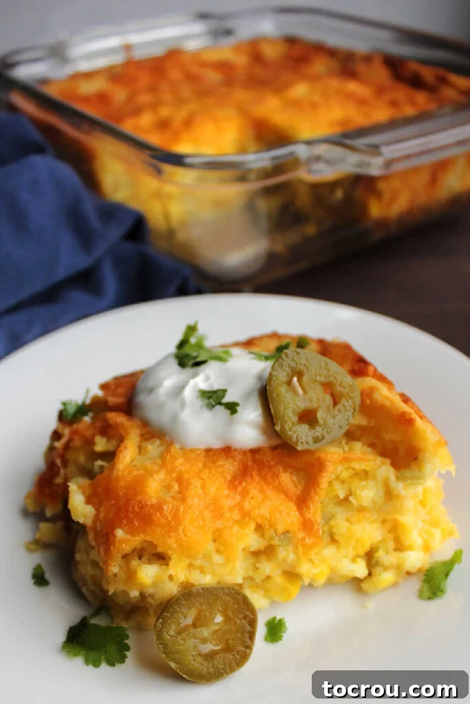 Individual Serving of Green Chile Corn Casserole Serving of corn casserole with green chiles and cheddar cheese topped with sour cream and jalapenos in front of pan.