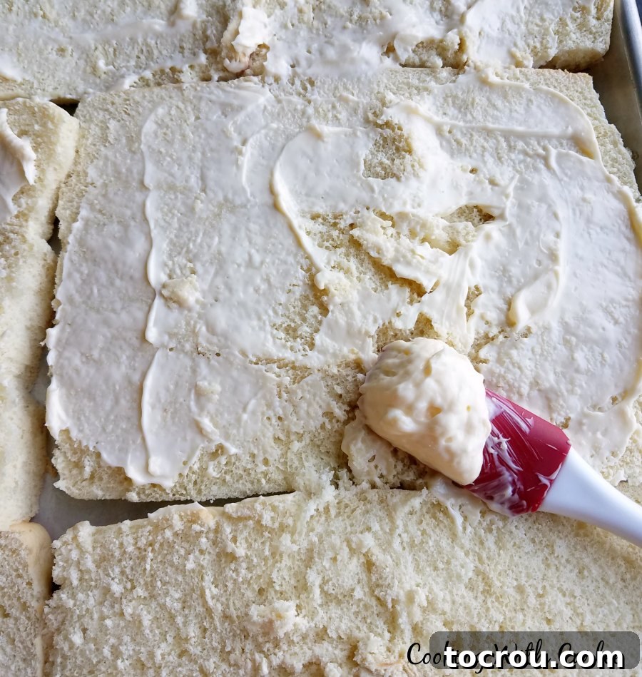 Spreading a creamy mayonnaise mixture evenly over the bottom halves of dinner rolls, preparing for layers of meat and cheese.