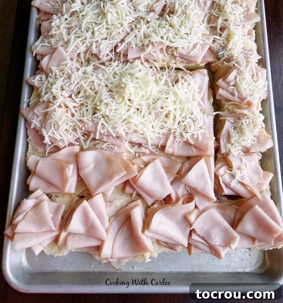 A tray of partially assembled turkey ham and cheese sliders, with layers of cheese, ham, and turkey waiting for the top buns.
