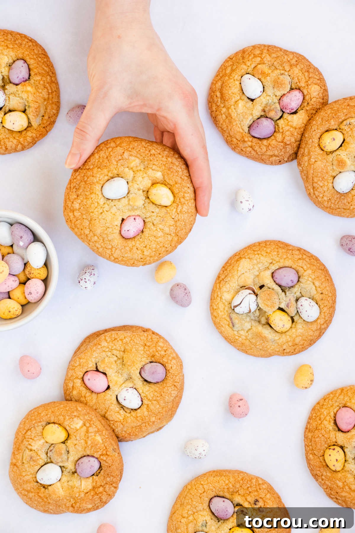 A hand gently holds a large, warm, golden brown cookie, freshly baked with melted mini chocolate eggs artfully pressed into its surface, ready for a delicious bite.
