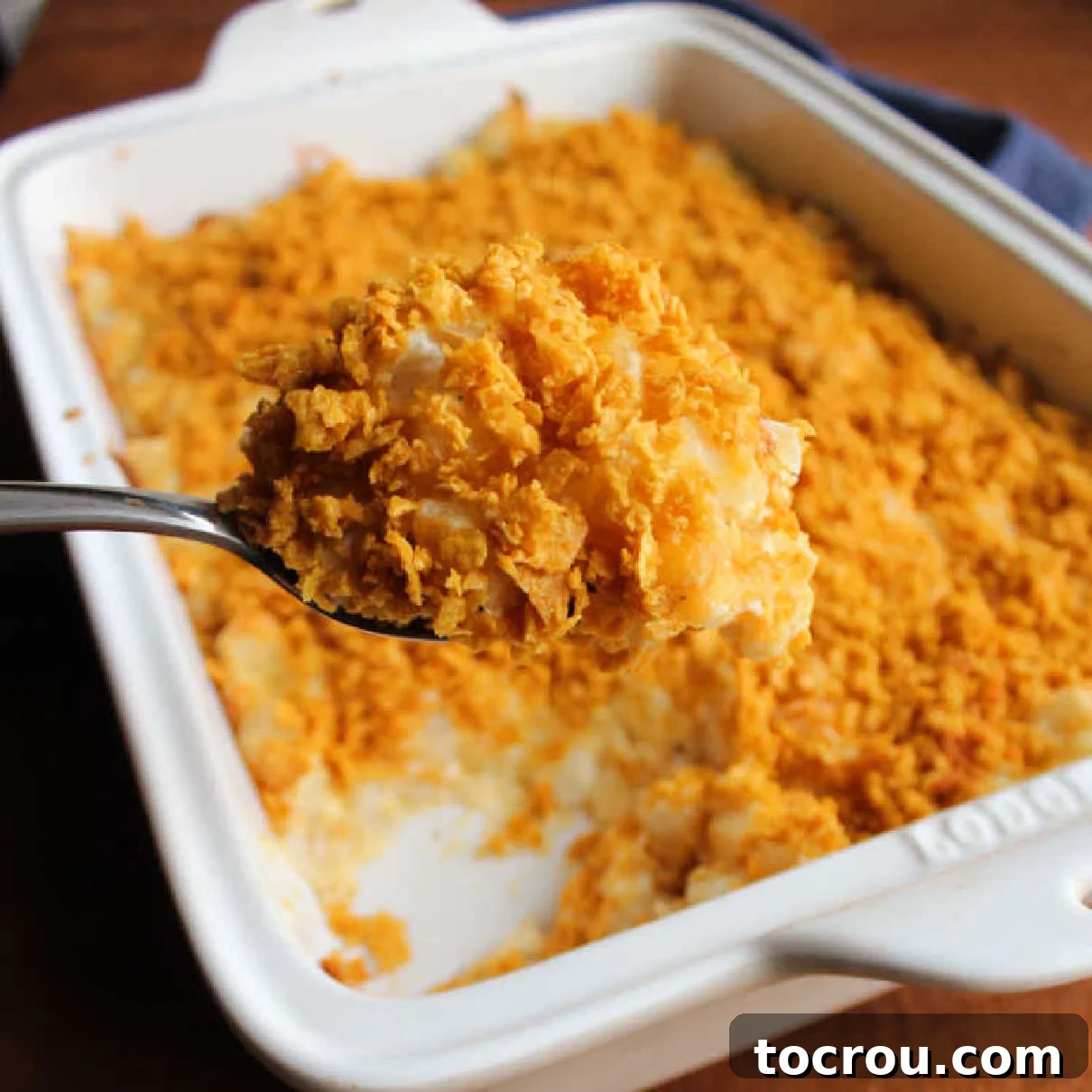Serving spoon getting a big scoop of French onion cheesy potato casserole topped with golden brown cornflake crumbs, indicating its creamy texture and delicious topping.