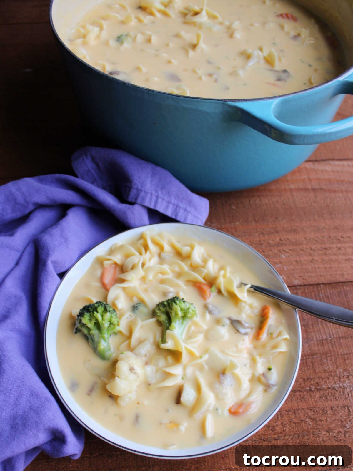 A cozy bowl of cheese soup, brimming with pasta, a colorful medley of vegetables, and soft potato chunks, sits invitingly beside a larger Dutch oven filled with more of the creamy Velveeta cheese soup.