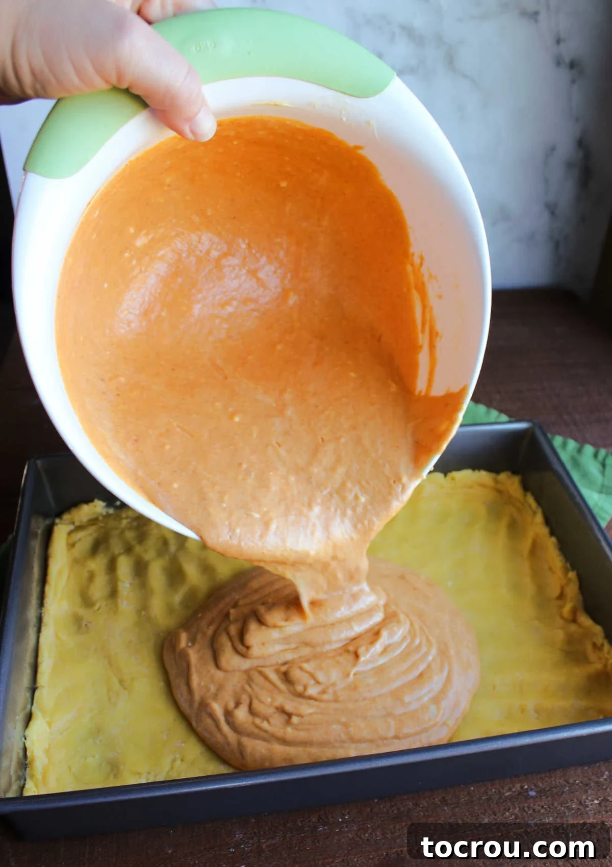 Ooey Gooey Pumpkin Butter Cake 7 A smooth, orange pumpkin cream cheese mixture being carefully poured from a mixing bowl over the pre-pressed cake mix crust in a baking pan, ready for the oven.