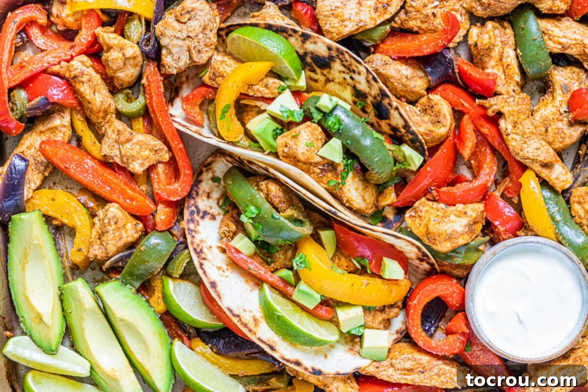 One-Pan Chicken Fajitas 2 Close up of chicken fajita mixture on sheet pan with colorful peppers, hunks of seasoned chicken, sliced avocado and more with two tortillas filled up and ready to eat.