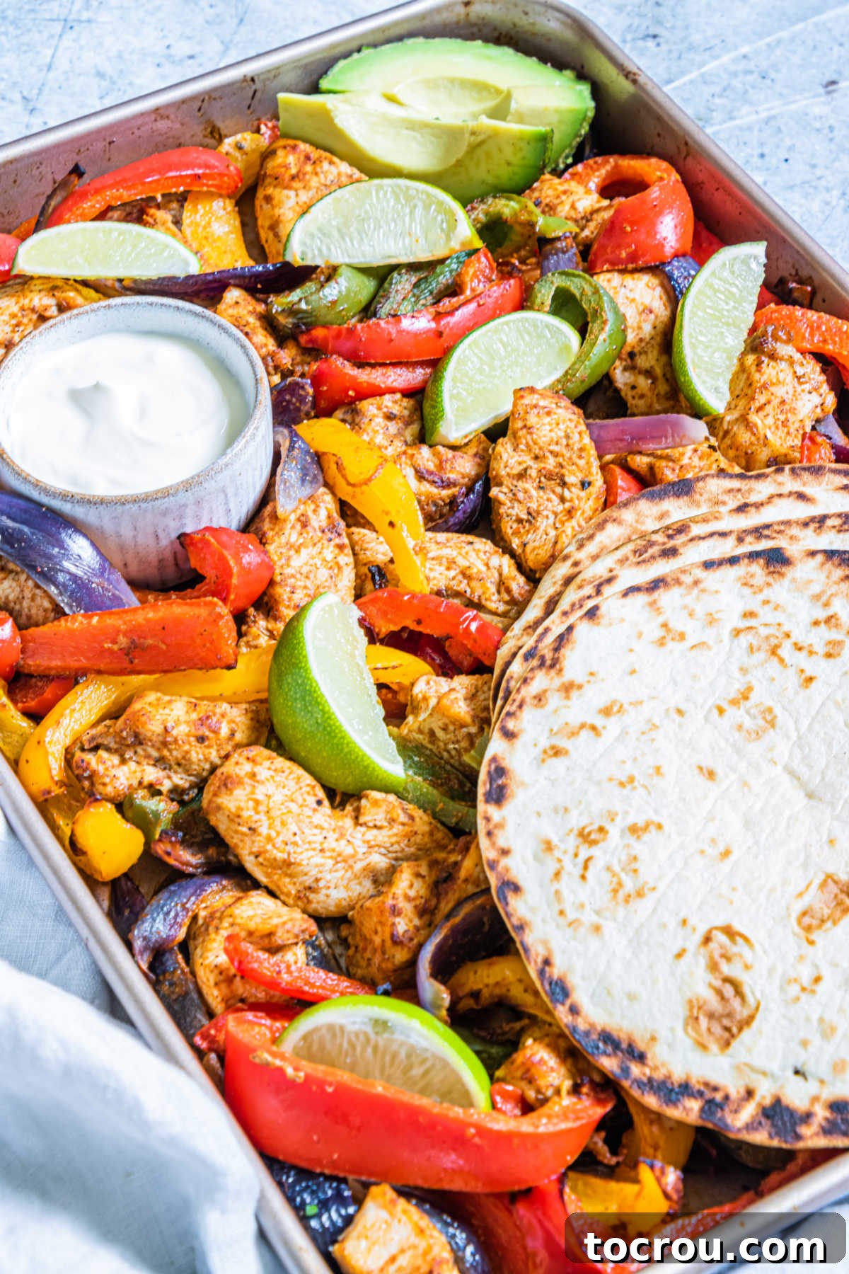 One-Pan Chicken Fajitas 3 sheet pan filled with chicken fajitas with warm tortillas, lime sedges and a ramekin of sour cream.