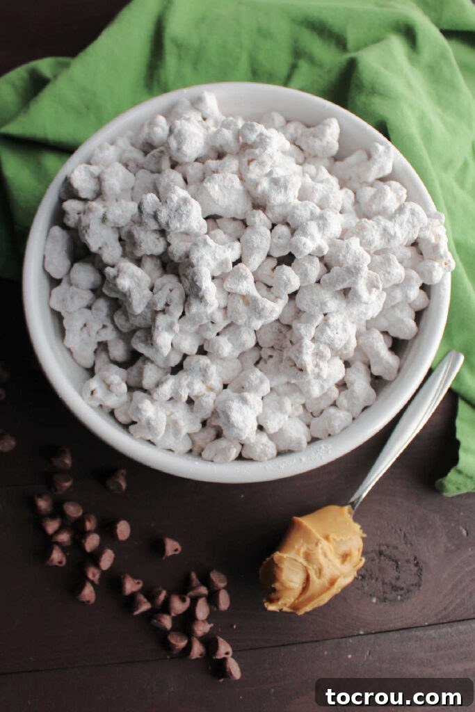 Ingredients for Puppy Chow Popcorn Popcorn coated in peanut butter, chocolate and powdered sugar in bowl next to spoonful of peanut butter and chocolate chips, highlighting ingredients.