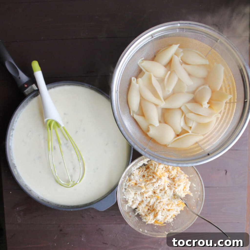 Ingredients for creamy chicken enchilada stuffed shells laid out: a bowl of shredded chicken and cheese, a pot of sour cream sauce, and a colander of cooked jumbo pasta shells.