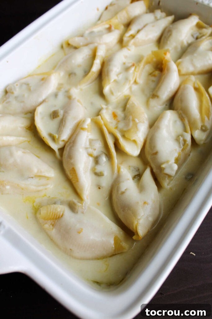 A baking dish filled with jumbo pasta shells, stuffed with chicken and generously covered in a rich white enchilada sauce, ready for the oven.