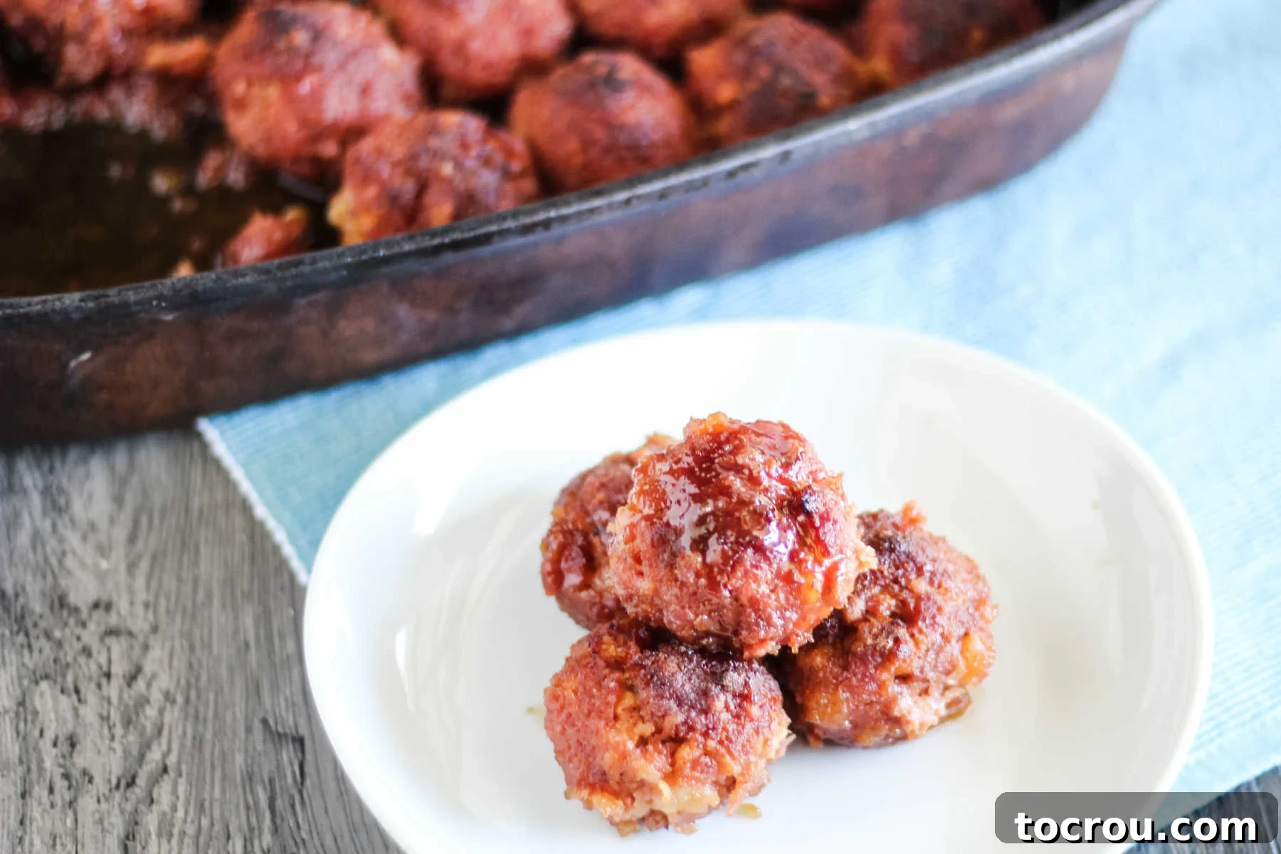 Plate of glazed ham balls with pan of remaining ham balls in the background, showcasing their delicious golden-brown glaze.