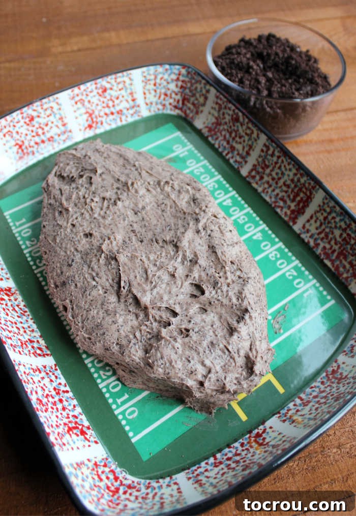 Oreo cheese ball mixture shaped like a football with bowl of Oreo crumbs nearby for coating the outside.