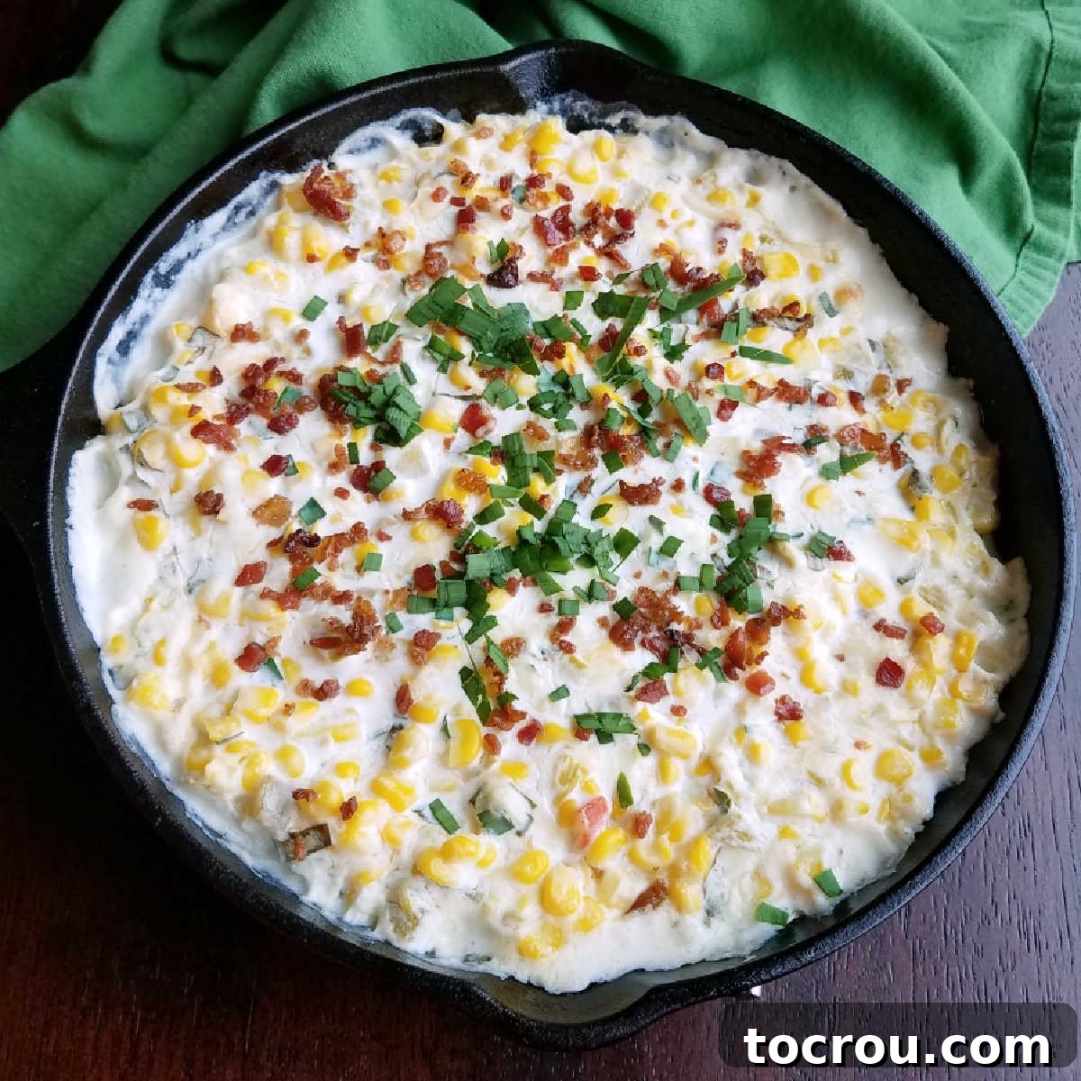 Ultimate Cheesy Corn Dip 2 A cast iron pan filled with golden, creamy corn dip, generously topped with crispy bacon bits, vibrant fresh cilantro, and chopped green onions, ready to be served.