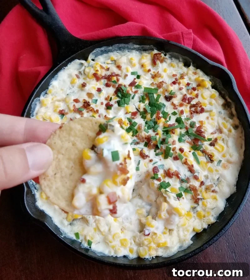 Ultimate Cheesy Corn Dip 6 A hand holding a tortilla chip generously loaded with creamy and spicy corn dip, showcasing the rich, melted cheese and speckled with herbs and bacon.