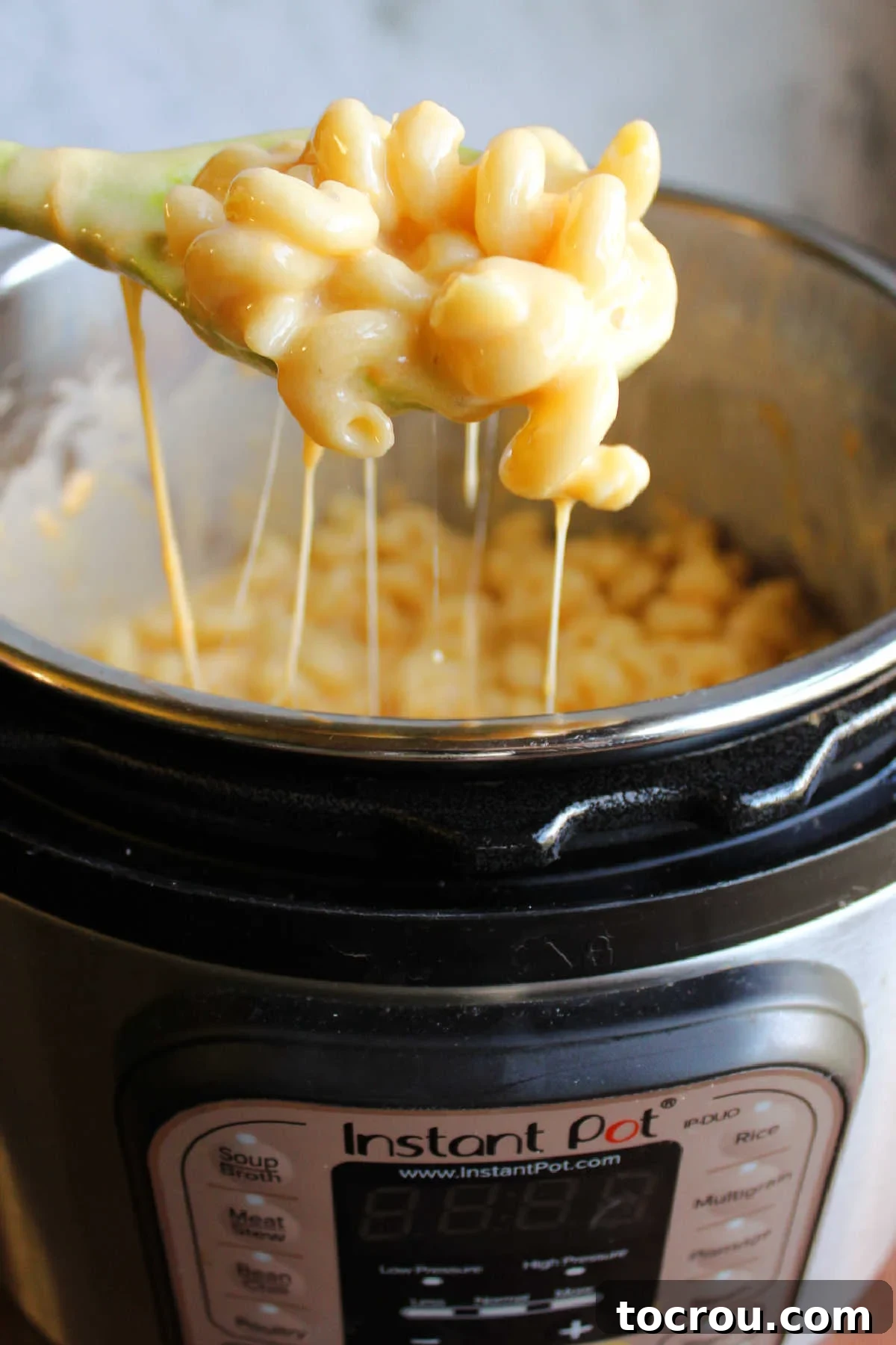 Spoon lifting curly mac and cheese instant pot with several cheese pulls coming from the pot showing rich, cheesy sauce.