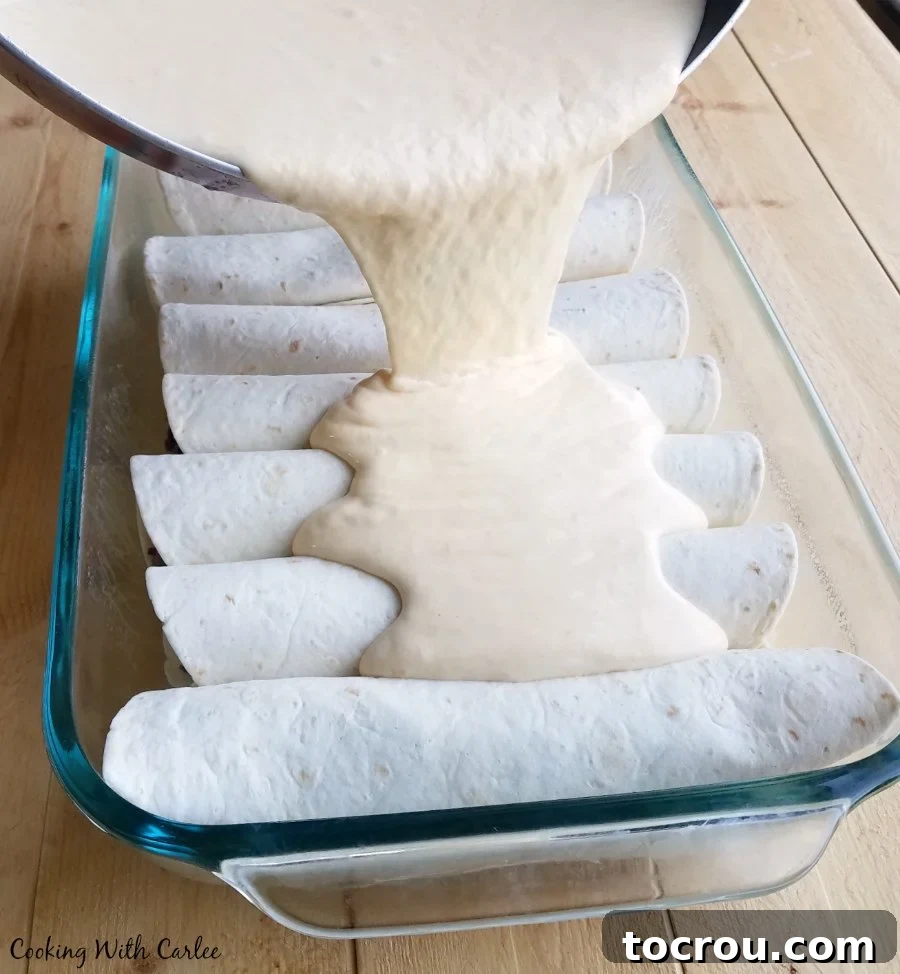 Pouring rich, creamy sour cream and cheese sauce generously over a pan of assembled enchiladas, ready for baking.