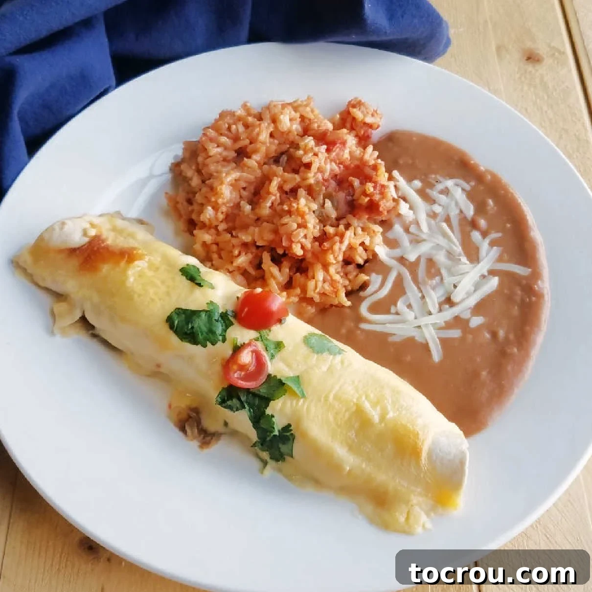 Creamy BBQ pulled pork enchilada, topped with fresh cilantro and diced tomatoes, served on a plate alongside refried beans and Spanish rice.