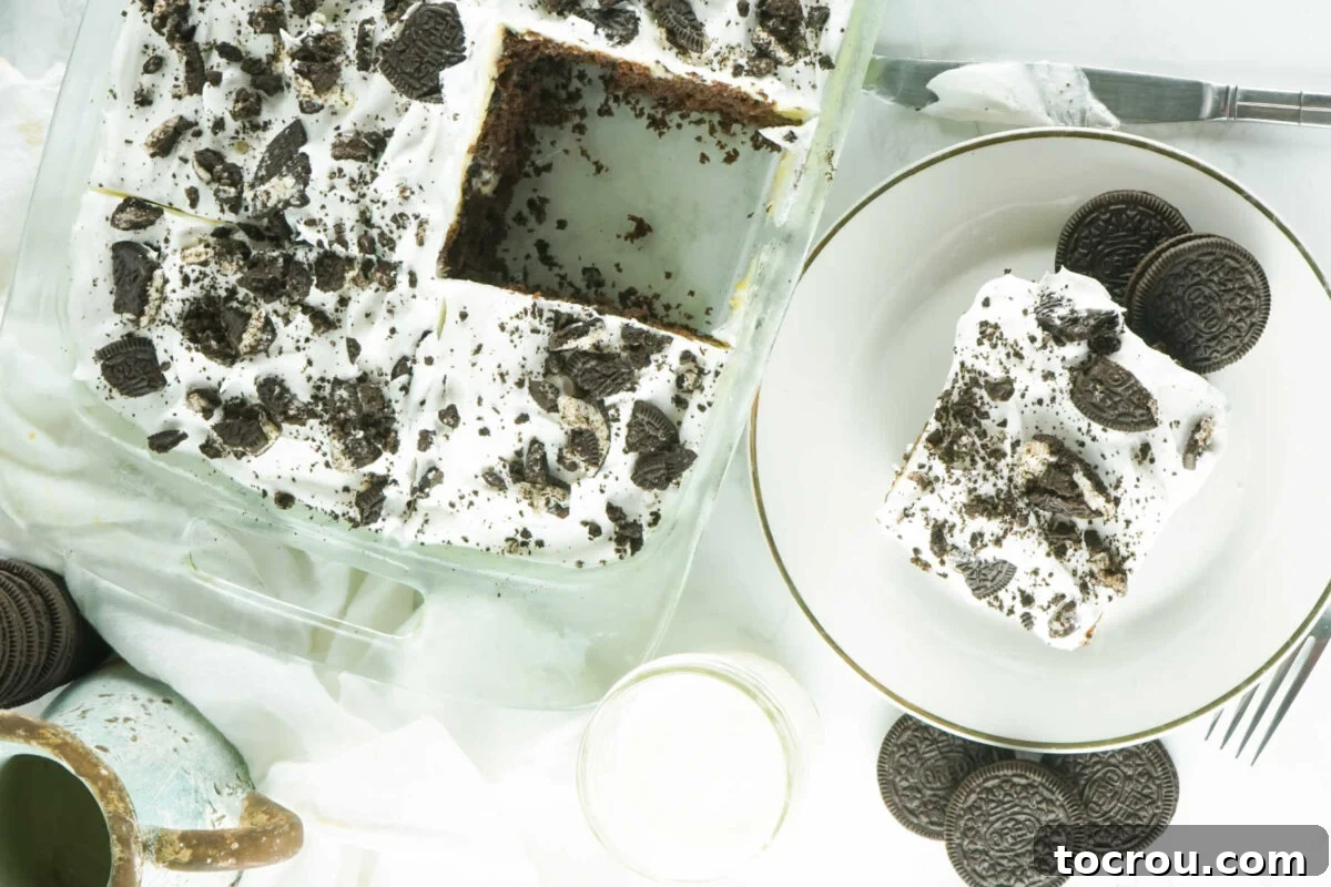 Hole-y Oreo Heaven 2 A 9x13 inch pan filled with a glorious chocolate cake, featuring creamy pudding infused within its poked holes, topped generously with fluffy whipped cream frosting and a scattering of crushed Oreos. One perfect slice has been removed and placed on a nearby plate, adorned with extra Oreo pieces.