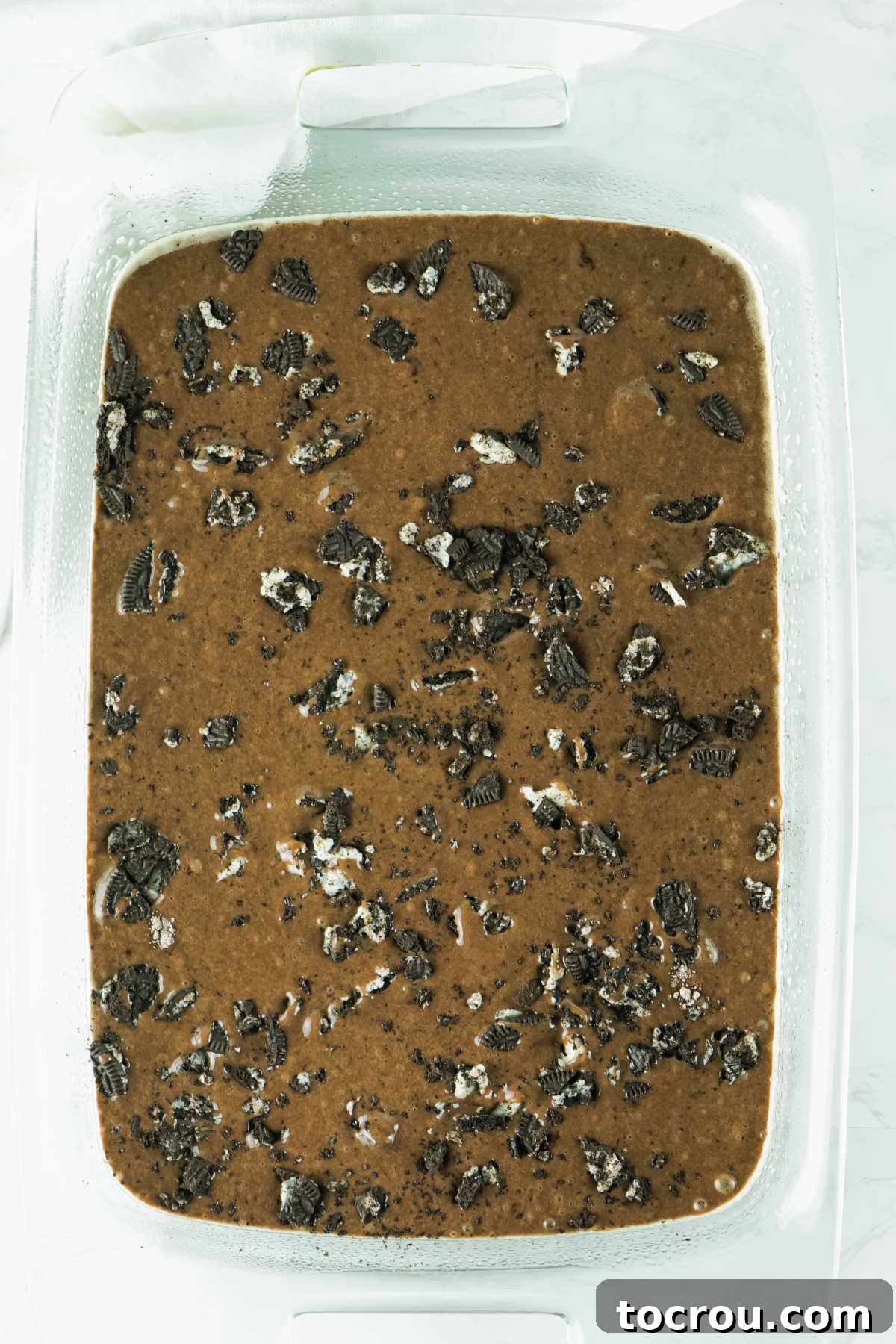 Hole-y Oreo Heaven 4 Chocolate cake batter, mixed to a smooth consistency and adorned with generously crushed Oreo pieces, spread evenly in a baking pan, ready for the oven to begin its transformation.