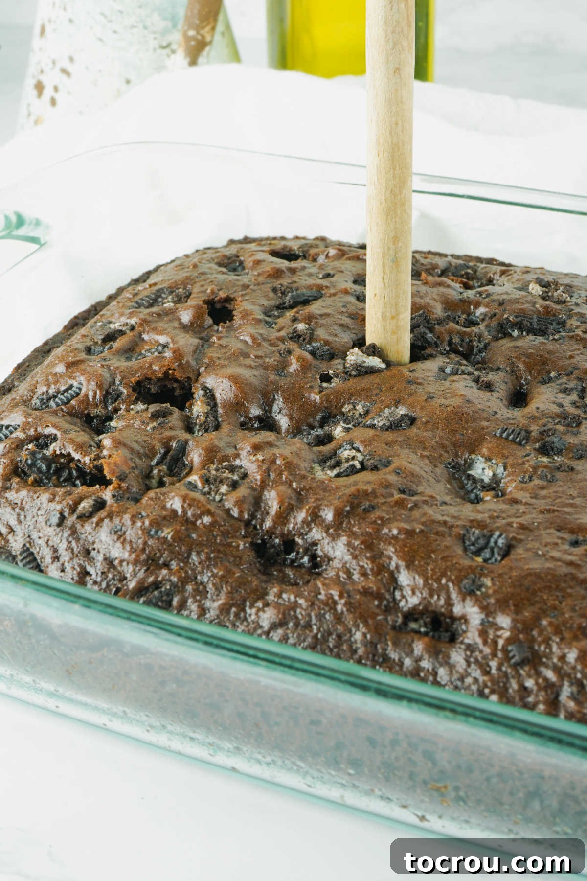 Hole-y Oreo Heaven 5 Close-up shot of a freshly baked chocolate cake, still warm, with numerous holes carefully poked across its surface using the handle of a wooden spoon, preparing it for the pudding infusion.
