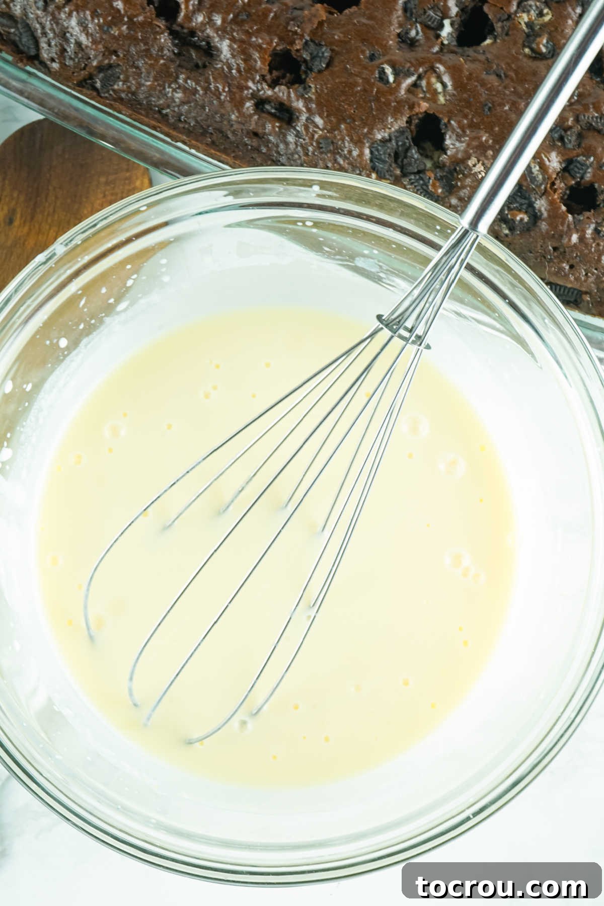 Hole-y Oreo Heaven 6 A mixing bowl containing instant pudding powder and fresh milk, being vigorously whisked together to create a smooth, creamy mixture, ready to be poured over the poked chocolate cake.