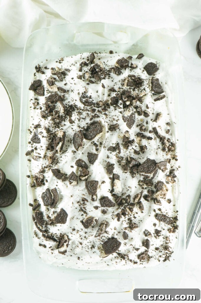 Hole-y Oreo Heaven 8 A complete Oreo poke cake, presented beautifully in a baking pan, topped with billowy white whipped cream and generously adorned with a scattering of dark, crushed Oreo cookie pieces, ready to be served.