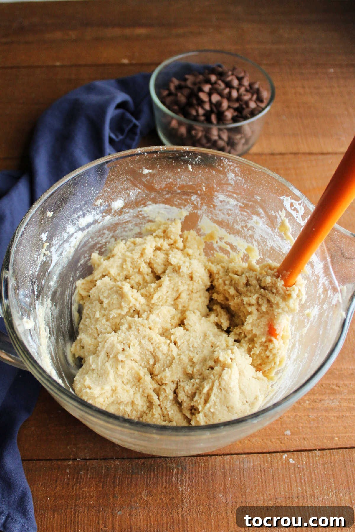 A glass mixing bowl filled with a creamy, light brown sugar and butter cookie dough. In the background, a small bowl holds chocolate chips.