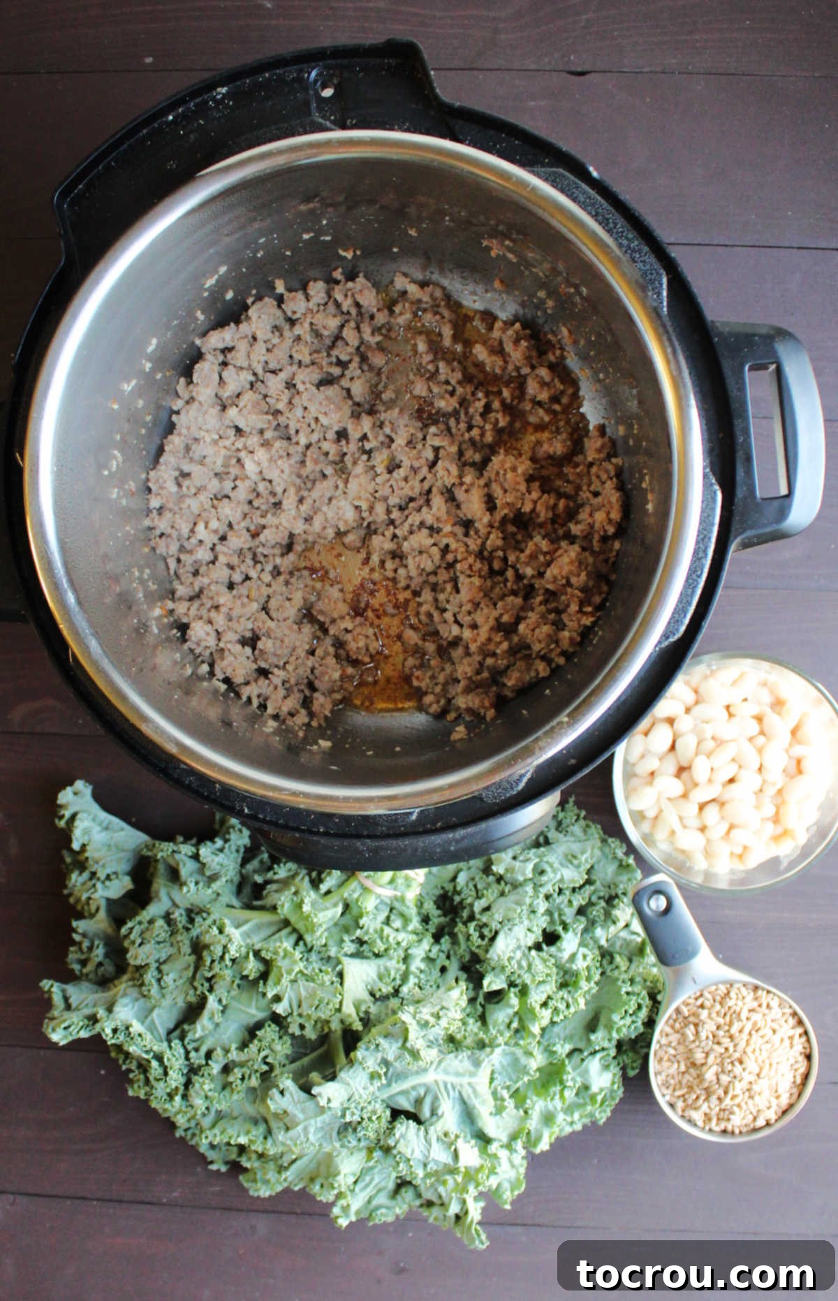 Browned Italian sausage sizzling in an Instant Pot, surrounded by fresh ingredients: a vibrant bunch of kale, a bowl of creamy white beans, and a cup of uncooked farro.