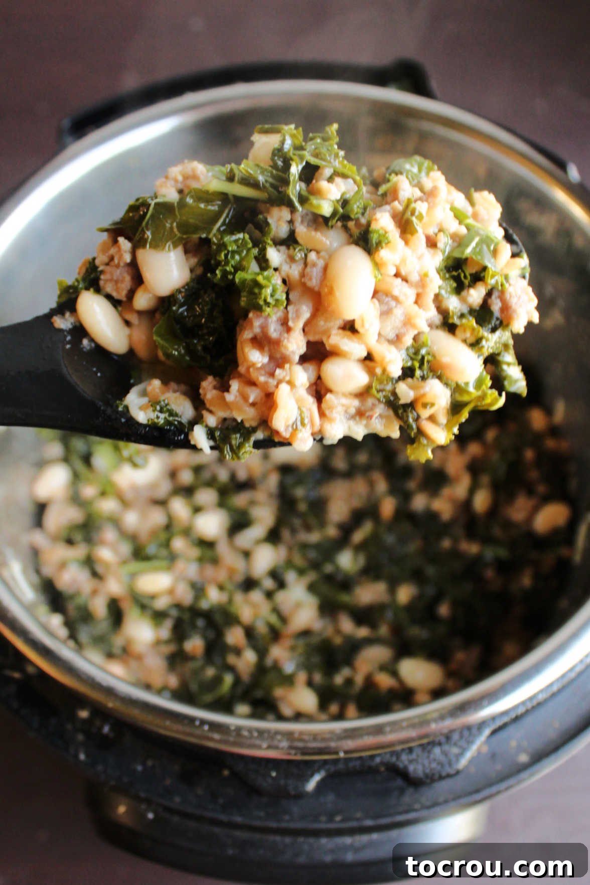 A spoonful of cooked farro, kale, and Italian sausage being lifted from the Instant Pot, showcasing the rich texture and steam of the freshly prepared meal.