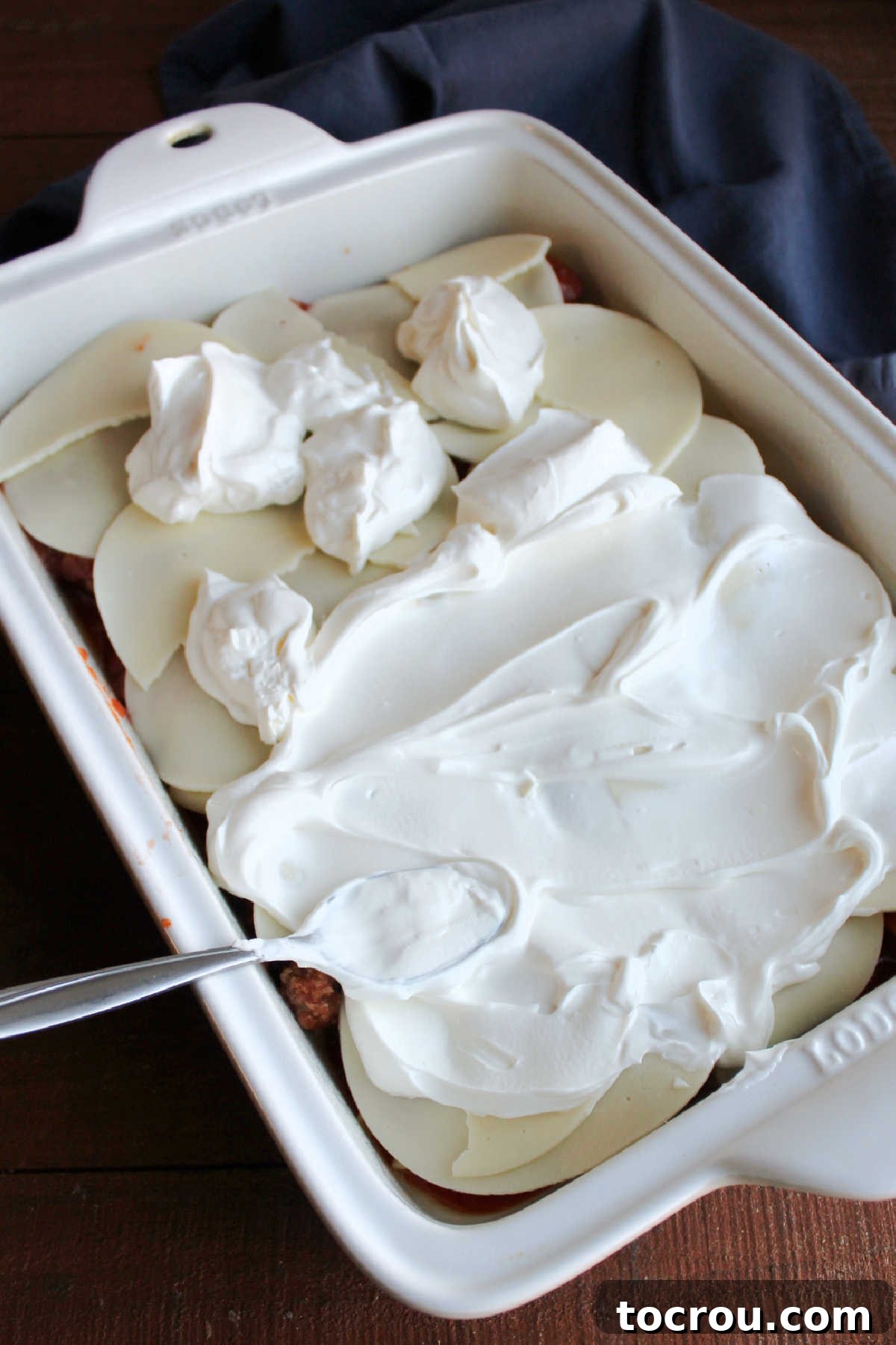 Layer of pasta and sauce topped with slices of provolone cheese, with a spoon spreading a creamy layer of sour cream over the cheese.