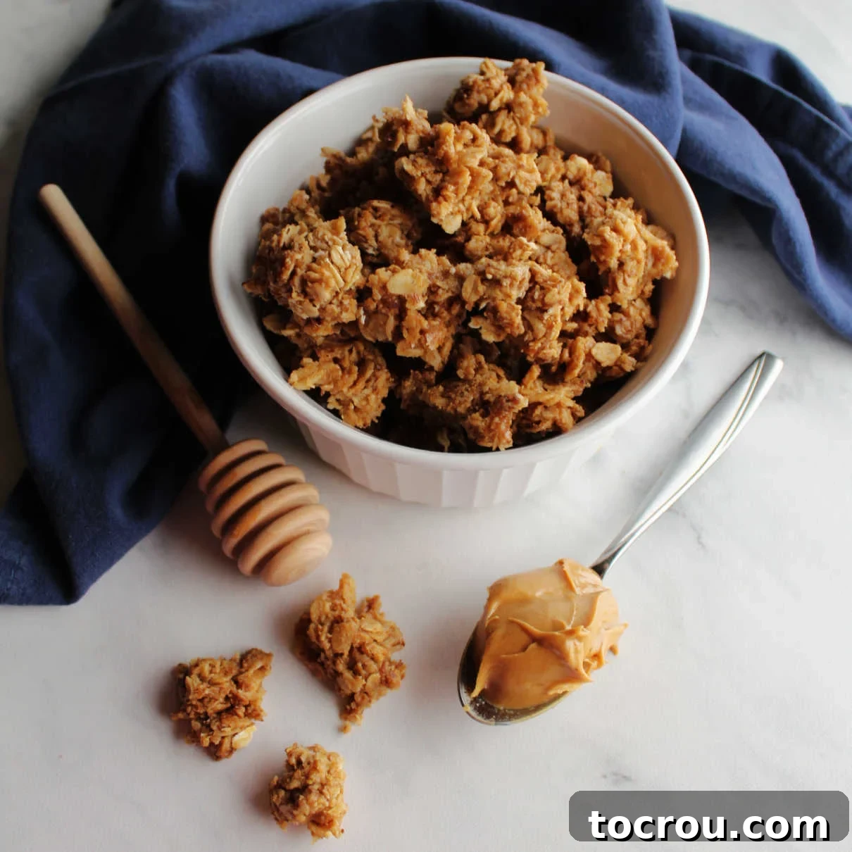 bowl of peanut butter granola next to a spoon of peanut butter and a honey drizzler.