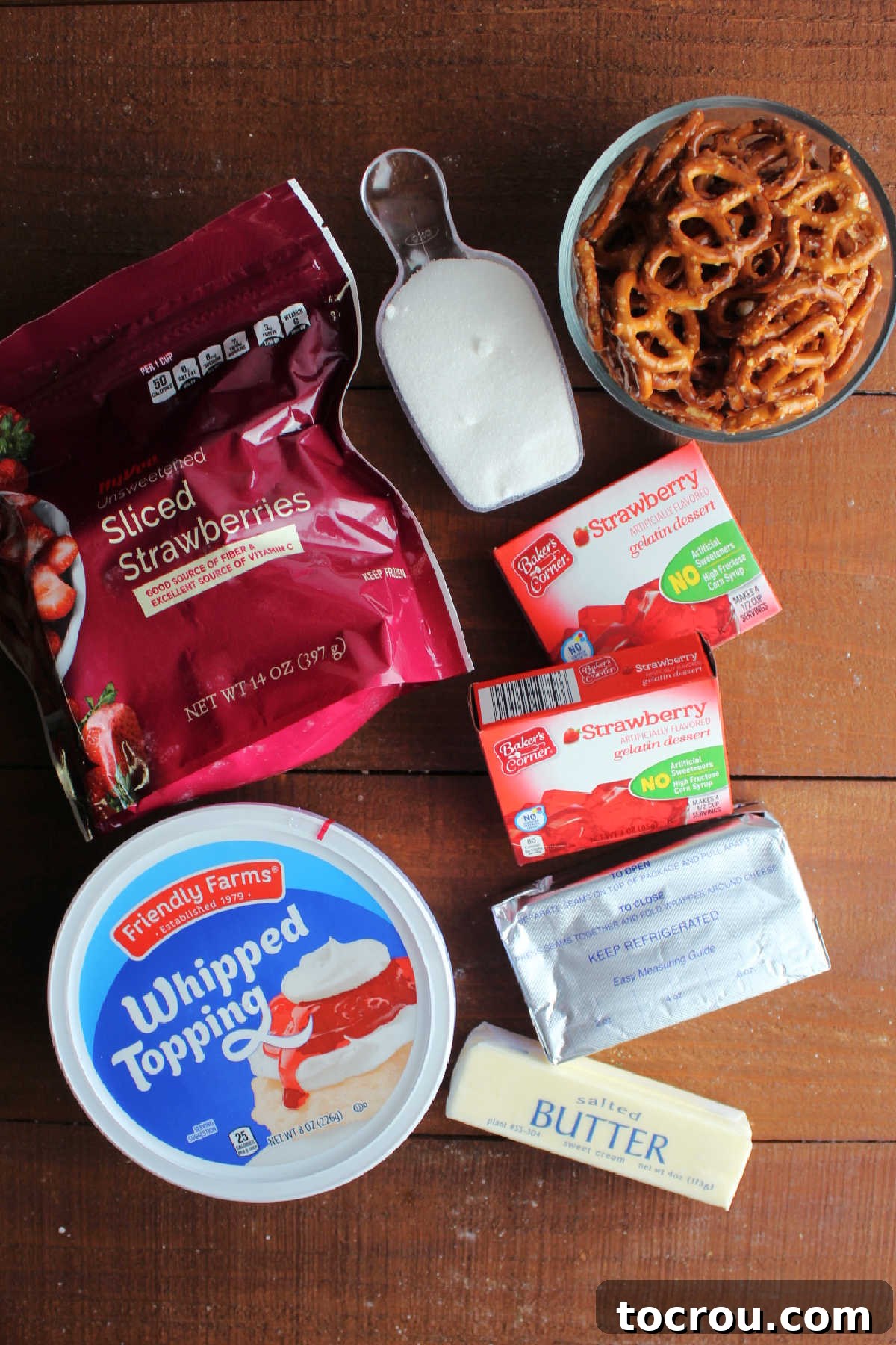 Strawberry Pretzel Salad Ingredients A flat lay of all the essential ingredients laid out for making Strawberry Pretzel Salad. Included are a bag of pretzels, granulated sugar, a stick of butter, a box of strawberry Jell-O, a bag of frozen sliced strawberries, and a tub of whipped topping, all perfectly arranged on a rustic wooden surface.