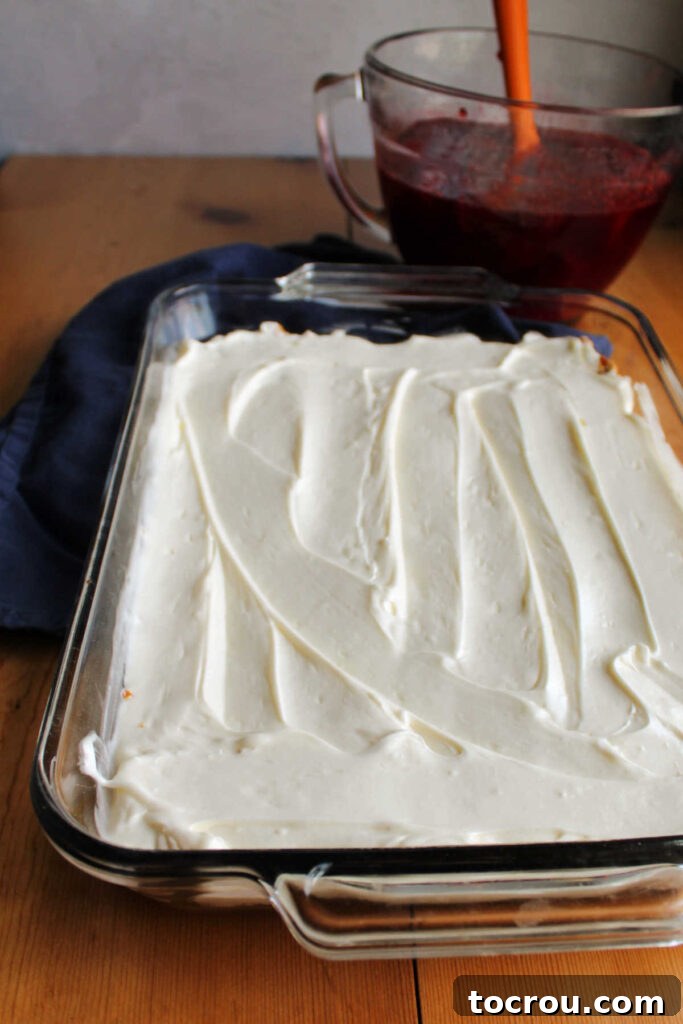 Cream Cheese Filling over Pretzel Crust The smooth, creamy cream cheese filling generously spread over the cooled pretzel crust, reaching all the way to the edges of the pan. In the background, a bowl of strawberry Jell-O mixture is visible, cooling down and preparing for the final layer.