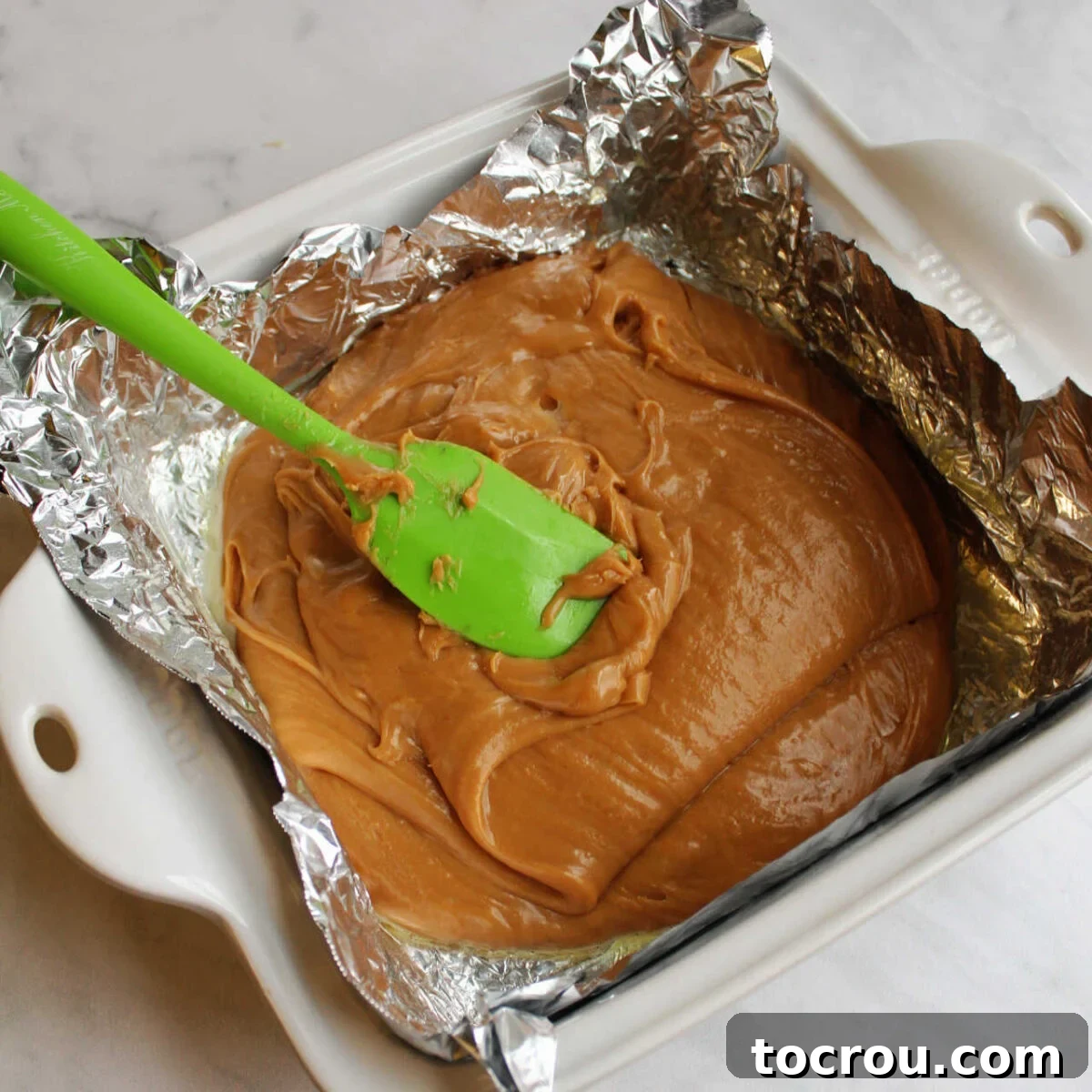 Fudge Mixture Poured into Pan Spreading smooth peanut butter fudge mixture into a foil-lined pan. The fudge is thick and uniform, being pressed evenly into the corners of the pan.