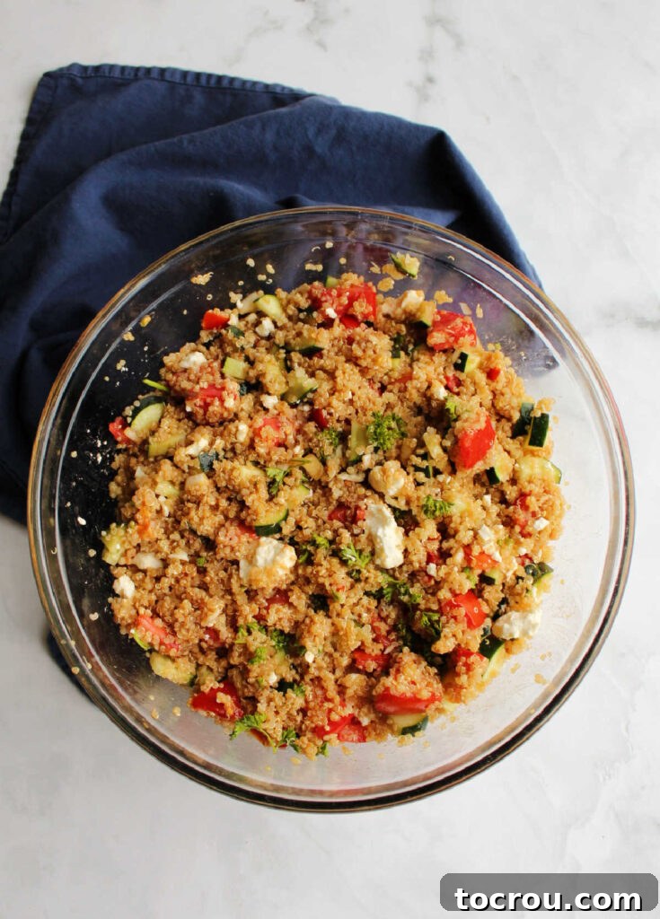 A large mixing bowl filled with a colorful medley of fresh ingredients including chopped tomatoes, crisp cucumbers, fluffy quinoa, dressing, crumbled feta cheese, and fragrant herbs, all ready for a final toss.