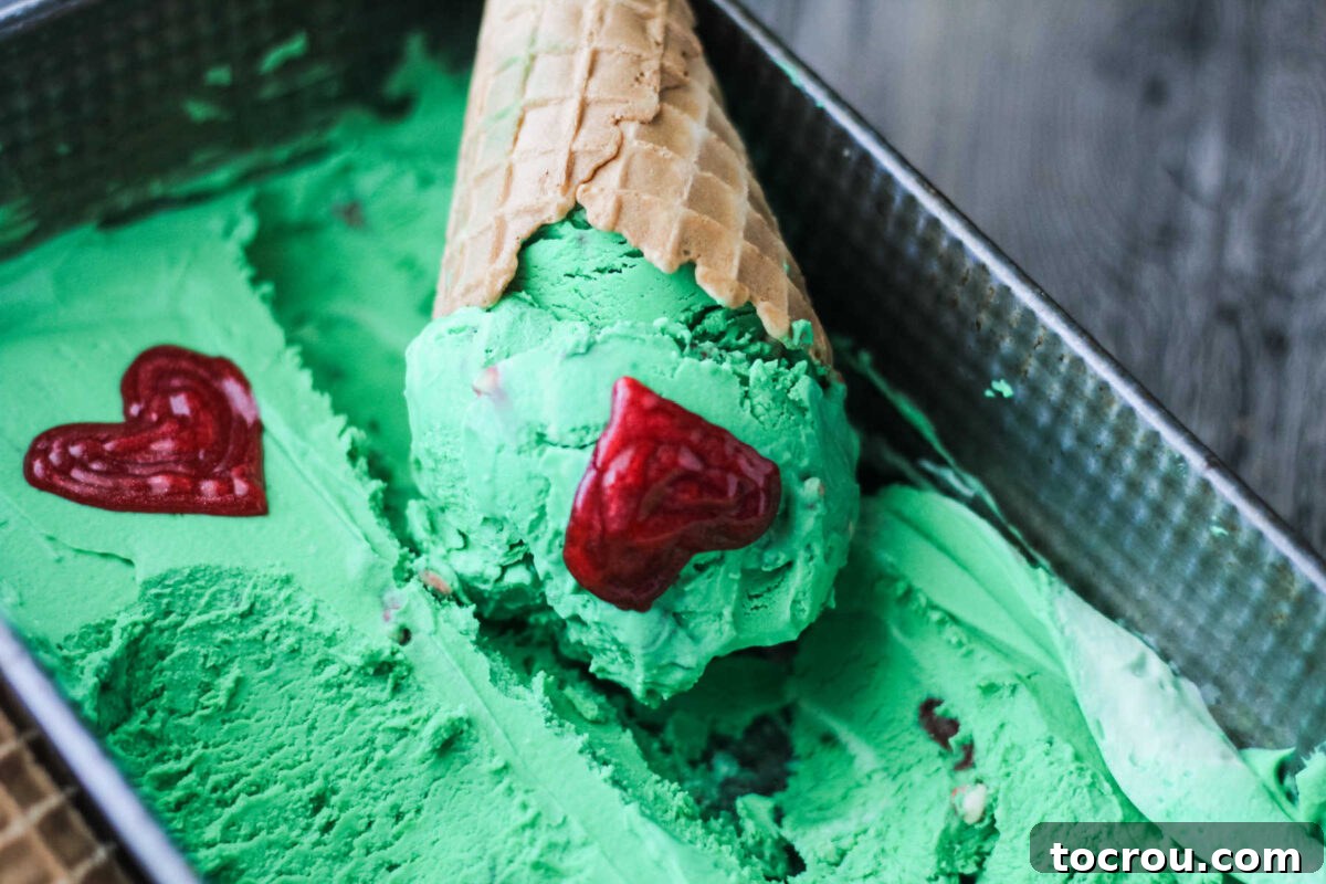 Grinch No-Churn White Chocolate Mint Ice Cream Green ice cream in a loaf pan with an ice cream cone topped with a scoop of it, decorated with red icing hearts to invoke the Grinch.