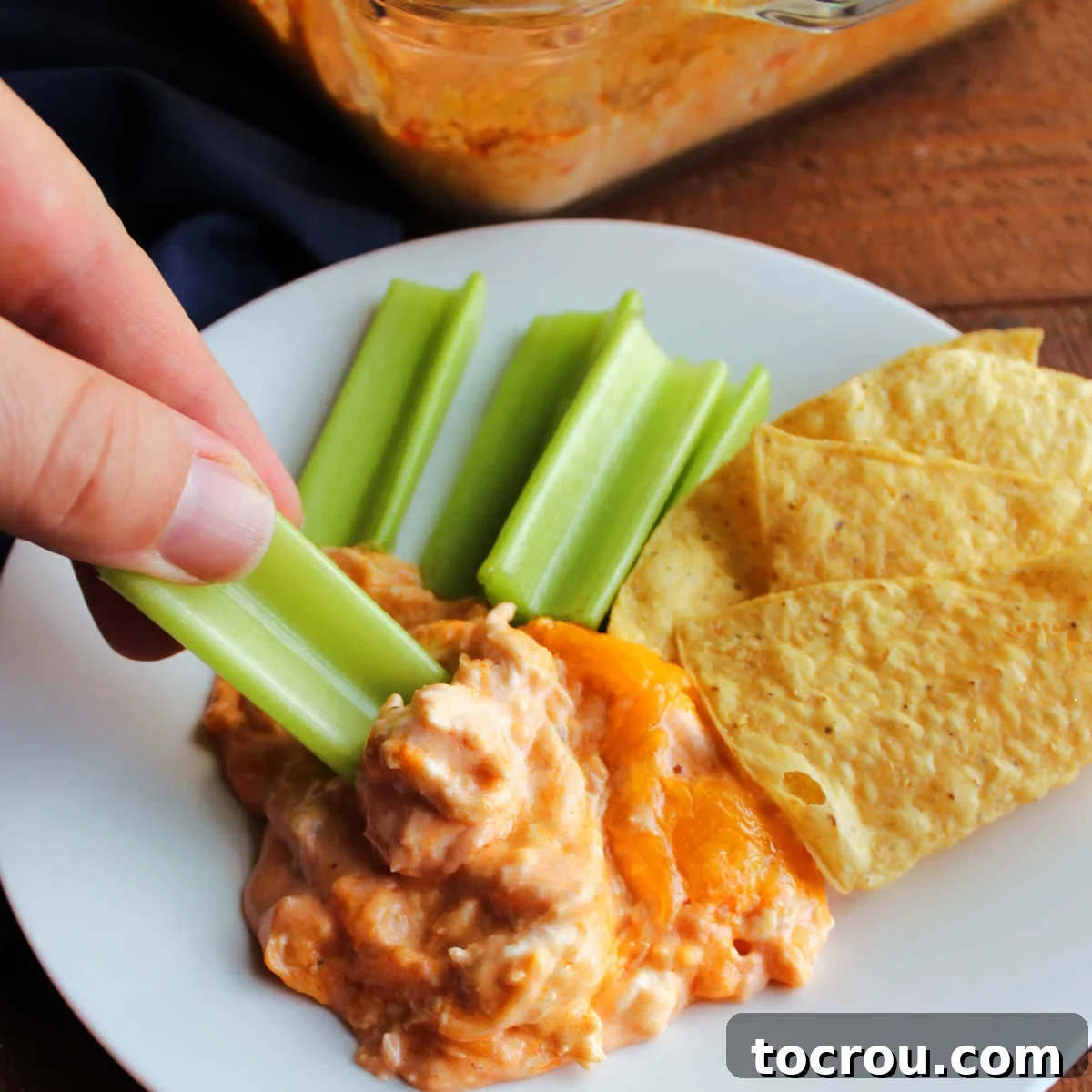 Ultimate Creamy Buffalo Chicken Dip 2 Hand dipping a fresh celery stick into warm, bubbling buffalo chicken dip, highlighting its creamy texture and the perfect golden-brown cheese topping.
