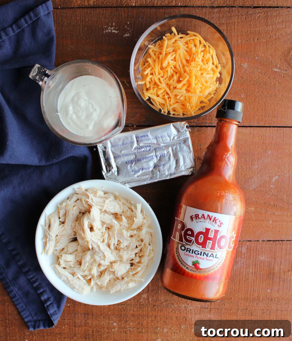 Ultimate Creamy Buffalo Chicken Dip 3 A vibrant flat lay of all the essential ingredients for buffalo chicken dip, including tender cooked chicken, a block of cream cheese, a bottle of ranch dressing, a mound of shredded cheddar cheese, and a classic bottle of Frank's hot sauce, all neatly arranged on a rustic wooden surface, ready for mixing.